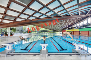 Bild vom Hallenbad mit drei Personen die jeweils von einem Startblock mit einem Köpfer ins Wasser springen. Mittig Diagonal ist ein roter Schriftzug mit Abgesagt